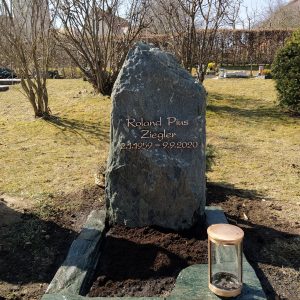 Dunkelgrüner Felsen-Grabstein in kompakter Naturform mit strukturierter Oberfläche und grüner Grabeinfassung