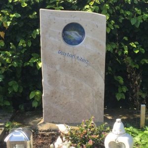 Kindergrabstein aus warmem Travertin mit runder Glaseinlage und blau-silbernem Engelsflügel-Motiv