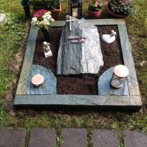 Urnenliegestein aus kleinem graugrünem Felsstein mit Kreuzgravur und dunkler Schieferplatteneinfassung